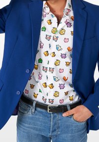 Blazer blu sopra una camicia bianca con personaggi colorati in pixel. Jeans di denim con una cintura nera intrecciata completano l'outfit.