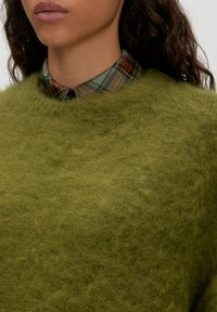 Olivgrüner, flauschiger Strickpullover mit weitem Rundhalsausschnitt, über ein karierstes Hemd in Grün-, Braun- und Marineblau-Tönen getragen.