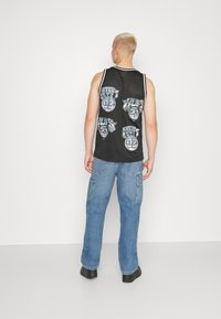 Camiseta sin mangas de malla negra con gráficos "HUFS" en gris. Combinada con unos pantalones cargo holgados de color azul claro y zapatos negros. Fondo minimalista.