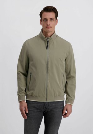 Man met kort donker haar, gekleed in een licht olijfgroene jas met rits en opstaande kraag, en donkere spijkerbroek, staand tegen een effen grijze achtergrond.