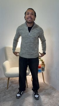 Pull-over gris clair texturé avec un col à demi-fermeture éclair, porté par-dessus une chemise noire, associé à un pantalon foncé et des baskets noires. Fond neutre.