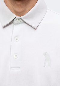 Gros plan d'une personne portant un polo blanc à trois boutons avec une petite figure en relief d'une personne sur la poitrine.