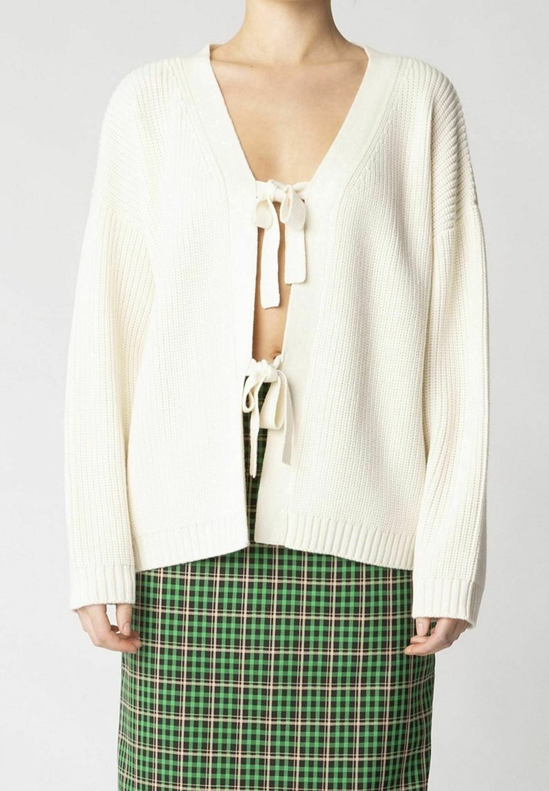 Femme portant un cardigan en maille crème noué avec des nœuds sur une jupe à carreaux verte et marine, présentée du cou jusqu’aux hanches.