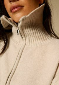 Suéter beige de cremallera con un alto cuello acanalado, que presenta una textura suave y sutiles detalles de costura a lo largo de la cremallera.