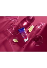 Un tubo di prodotto per la cura della pelle con una sfumatura viola e blu, centrato, circondato da contenitori di vetro trasparente su una superficie rosa, con fette di limone.