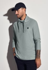 Světlezelený texturovaný pulovr s dlouhým rukávem a rolákem, na hrudi s černým kruhovým logem, nošený s tmavými kalhotami a černou kšiltovkou.