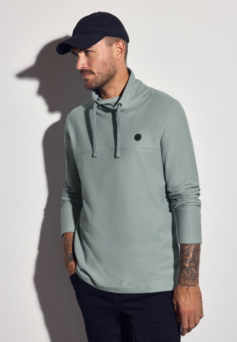 Světlezelený texturovaný pulovr s dlouhým rukávem a rolákem, na hrudi s černým kruhovým logem, nošený s tmavými kalhotami a černou kšiltovkou.