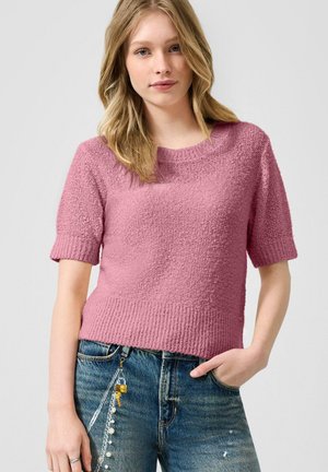 Jeune femme portant un pull rose texturé à manches courtes et un jean bleu avec un porte-clés accroché à la boucle de ceinture.