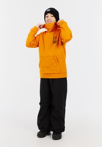 Oranžová mikina s vysokým golierom a predným vreckom, zdobená grafickým potlačou. V kombinácii s čiernymi, voľnými nohavicami a čiernymi čižmami.