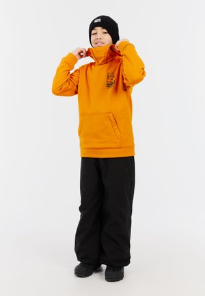 Oranje hoodie met een hoge kraag en een voorzak, voorzien van een grafische print. Combinatie met zwarte, loszittende broek en zwarte laarzen.