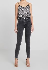 Femme portant un débardeur à motifs noir et blanc, un jean skinny noir délavé taille haute, et des sandales à talons beiges, se tenant sur un sol gris.