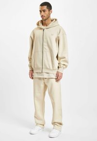 Beige Kapuzenpullover und passende Hose aus weichem Stoff; verfügt über einen Reißverschluss vorne, große Taschen und eine lockere Passform mit minimaler Markenführung. Weiße Sneaker.