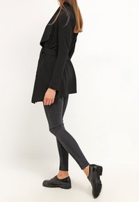 Musta kääritty takki, jossa on laskostettu etuosa, vyötäröllä vyö, yhdistetty tummanharmaisiin kapeisiin farkkuihin ja mustiin nauhakenkiin, joissa on rakenteellinen pohja.