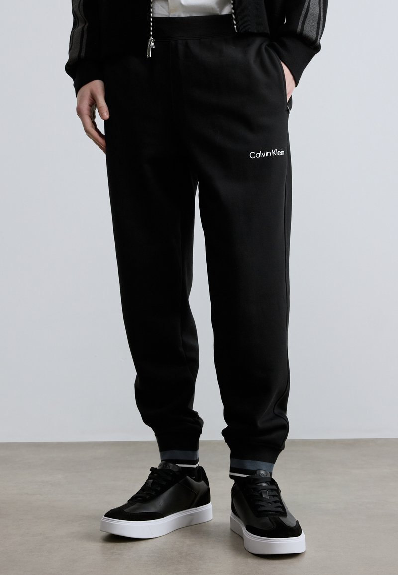 Calças de treino pretas com cintura elástica, apresentando bolsos laterais e um logótipo branco da "Calvin Klein". Combinadas com sapatilhas pretas e brancas.