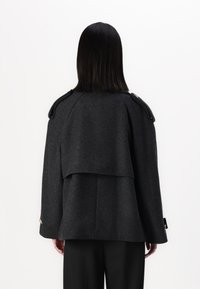 Manteau en mélange de laine gris avec un dos superposé, des détails d'épaule marquants et des accents dorés aux poignets. Présente une silhouette ample et structurée.