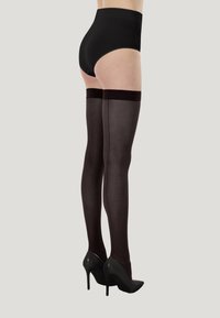 Culotte noire taille haute associée à des bas résille noirs transparents montant jusqu'en haut des cuisses. Le mannequin porte des escarpins noirs, mettant en avant un design élégant.