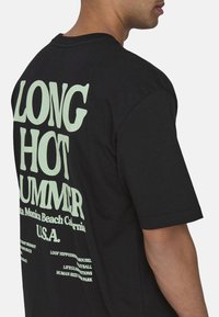 Czarny bawełniany t-shirt z dużym, jasnoczonym tekstem z tyłu. Tekst zawiera "DŁUGIE GORĄCE LATO" oraz szczegóły lokalizacji w swobodnej czcionce.