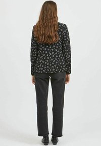 Langärmlige schwarze Bluse mit weißem Blumenmuster, kombiniert mit dunkelgrauen, geraden Hosen und schwarzen Schuhen, von hinten betrachtet.