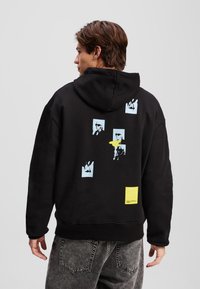 Fekete kapucnis pulóver, melynek hátulján több kék grafikai nyomat található, az alján egy sárga kiemelő négyzettel. Puha szövet textúra.