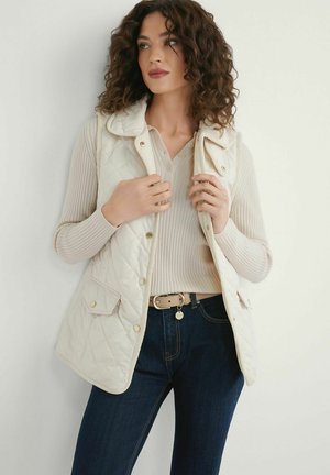 Kobieta o kręconych włosach nosząca beżową pikowaną kamizelkę na prążkowanym swetrze, ciemne dżinsy i beżowy pasek, patrząca na bok na białą ścianę.