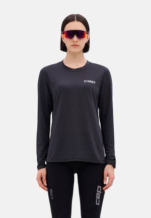 Woman wearing black long-sleeve athletic shirt and leggings with gray "cep" logos, and reflective sports sunglasses, standing against a plain background.