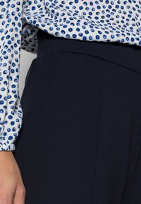 Halenka s dlouhým rukávem s bílým podkladem a modrým puntíkovaným vzorem, která je spárována s vysokými navy kalhotami s pevnou texturou.