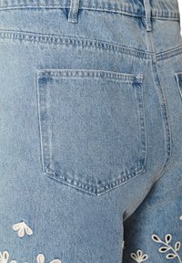 Φωτεινό μπλε τζιν παντελόνι με πίσω τσέπη και λευκά κεντημένα λουλουδάτα σχέδια κοντά στην περιοχή του ισχίου και του μηρού.