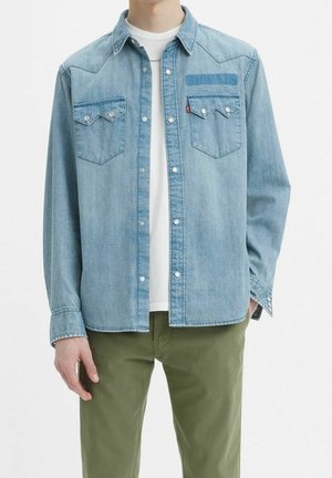 Persoon draagt een lichtblauw denim overhemd over een wit T-shirt en olijfgroene broek, staat met één hand gedeeltelijk in de zak.