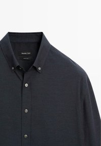 Camicia a maniche lunghe blu scuro con colletto a punta, caratterizzata da una texture sottile e quattro bottoni lungo la chiusura frontale. Etichetta del marchio visibile.