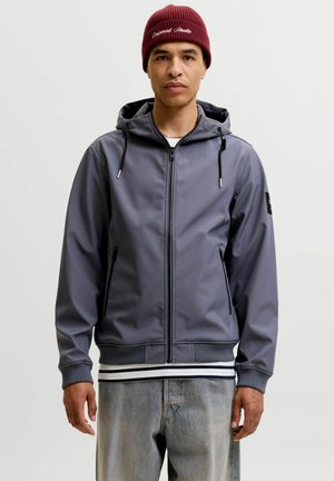 Young man wearing a maroon beanie and zipped gray hooded jacket over a striped shirt and light blue jeans standing against a plain background.