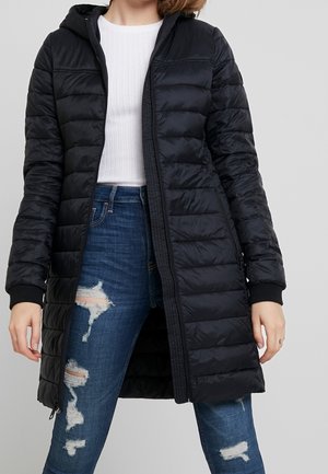 Femme portant une longue doudoune noire matelassée sur un haut blanc côtelé et un jean bleu déchiré, debout devant un fond gris uni.