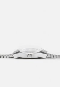Reloj de acero inoxidable con una caja redonda, correa de malla gris y una cara de vidrio liso. Presenta una corona prominente en el lateral.