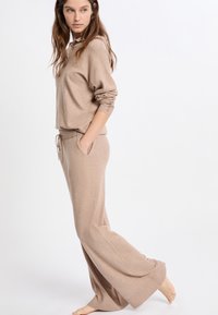 Lichtbruine loungewear set bestaat uit een gebreid hoodie en wijde broek. Kenmerken zijn een trekkoord in de taille, zijzakken en geribbelde manchetten.