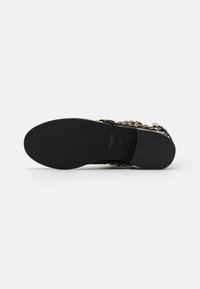 Zapato negro con una suela lisa y plana que cuenta con un acento de cadena dorada. La parte superior tiene un diseño de tiras y un acabado sintético texturizado.