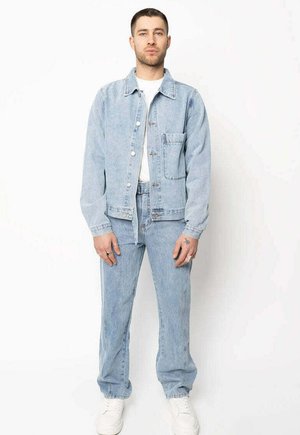 Homme debout face à l'objectif, portant une veste en jean bleu clair et un jean avec des baskets blanches et une chemise blanche sur un fond uni.