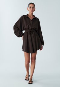 Rjava srajčna obleka s sponkami na gumbke, z zateznim pasom, dolgimi napihnjenimi rokavi in razširjenim robom, kombinirana s črnimi sandali s paščki.