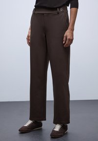 Pantalons larges marron en tissu doux, avec une taille plate et des boutons, des poches latérales et une coupe décontractée. Portés avec des baskets sombres.