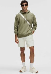 Haut à capuche vert olive avec cordons, shorts crème avec poches, et baskets neutres. Le modèle porte des lunettes de soleil et des chaussettes blanches.