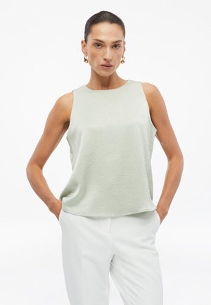 Femme aux cheveux foncés portant un haut sans manches clair et texturé et un pantalon blanc, posant avec les mains dans les poches sur un fond blanc.