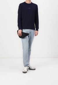 Chemise à manches longues bleu marine avec logo GAP, jean bleu clair et sac banane noir à la main ; chaussures de sport blanches et grises ; arrière-plan minimal.