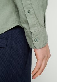 Hellgrünes Langarmhemd mit Knopfmanschetten, strukturiertem Stoff und sichtbaren Nähten. Dunkelblaue Taschenapplikation der dazugehörigen Hose.