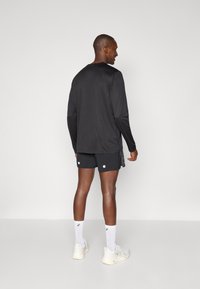 Black long-sleeve shirt paired with black shorts featuring two white dots; model wears white sneakers and ankle-length socks.