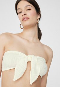 Žlutý bezramínkový bikini top s uzlem v přední části, vyrobený z texturované látky a jemnými horizontálními pruhy.