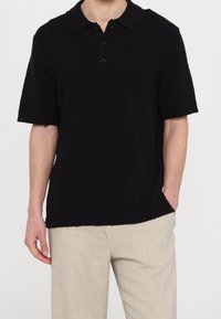 Mies, który ma na sobie czarną koszulę polo z krótkim rękawem i jasnobeżowe, teksturowane spodnie, z jedną ręką w kieszeni na jednolitym tle.