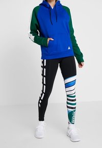 Blå huvtröja med gröna och vita ärmar; svarta leggings med flerfärgade geometriska mönster. Vita sneakers fulländar outfiten.