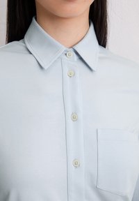Hellblaues Button-Up-Hemd mit spitzem Kragen, vier Knöpfen, kleiner Brusttasche und glatter Stoffstruktur.