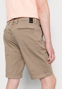 Shorts in tessuto beige con un dettaglio della tasca con zipper sul retro e un orlo piegato, con un'etichetta nera del brand in vita.