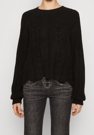 Pull noir en maille torsadée à manches longues associé à un jean gris à lacets sur le devant, porté par une personne debout.