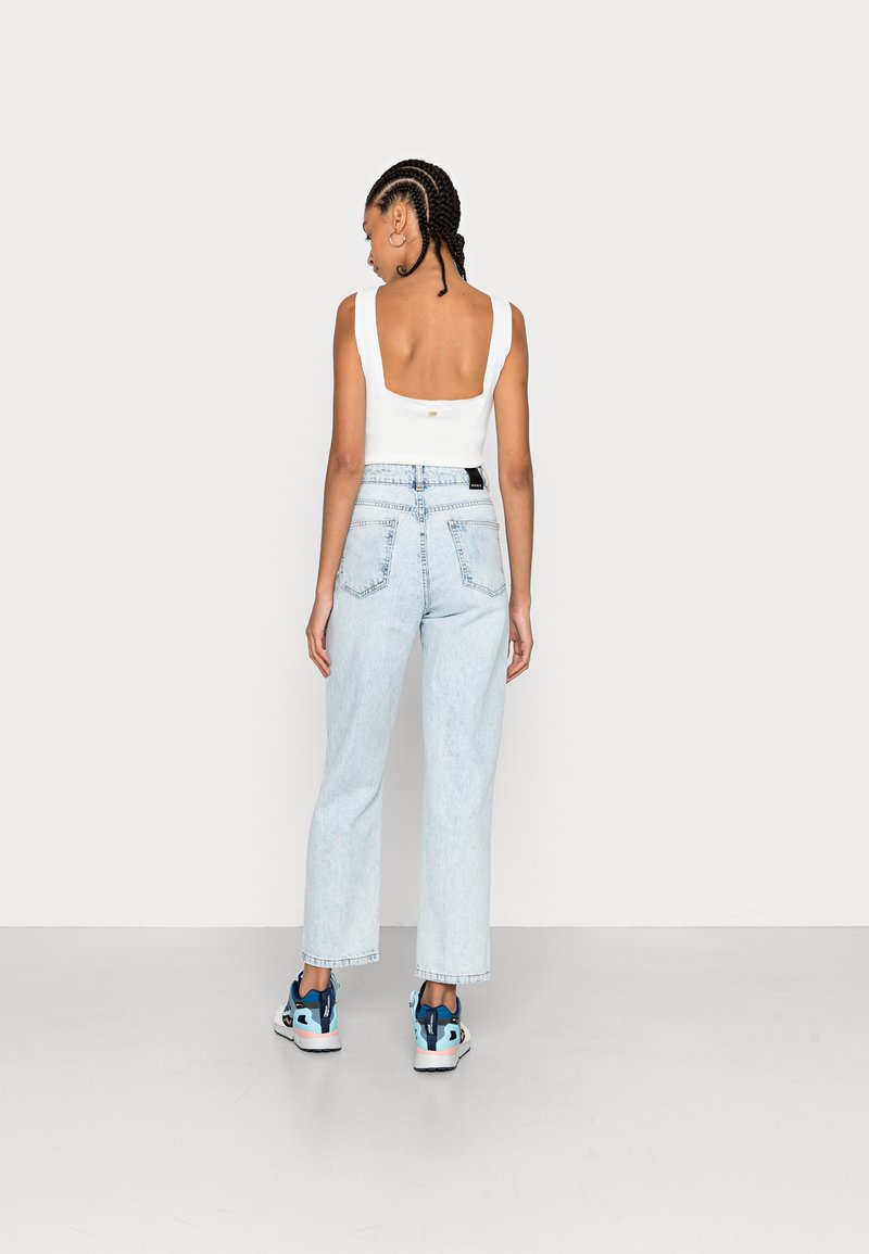 Weißes ärmelloses Top mit quadratischem Rückenteil, kombiniert mit hellblauen hochgeschnittenen Jeans und Sneakers in Blau, Rosa und Schwarz. Einfarbiger Hintergrund.