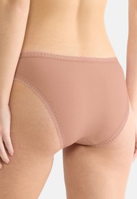 Set bagfra af person iført højtaljede malva undertøj med blondeckant på talje og benåbninger mod hvid baggrund.